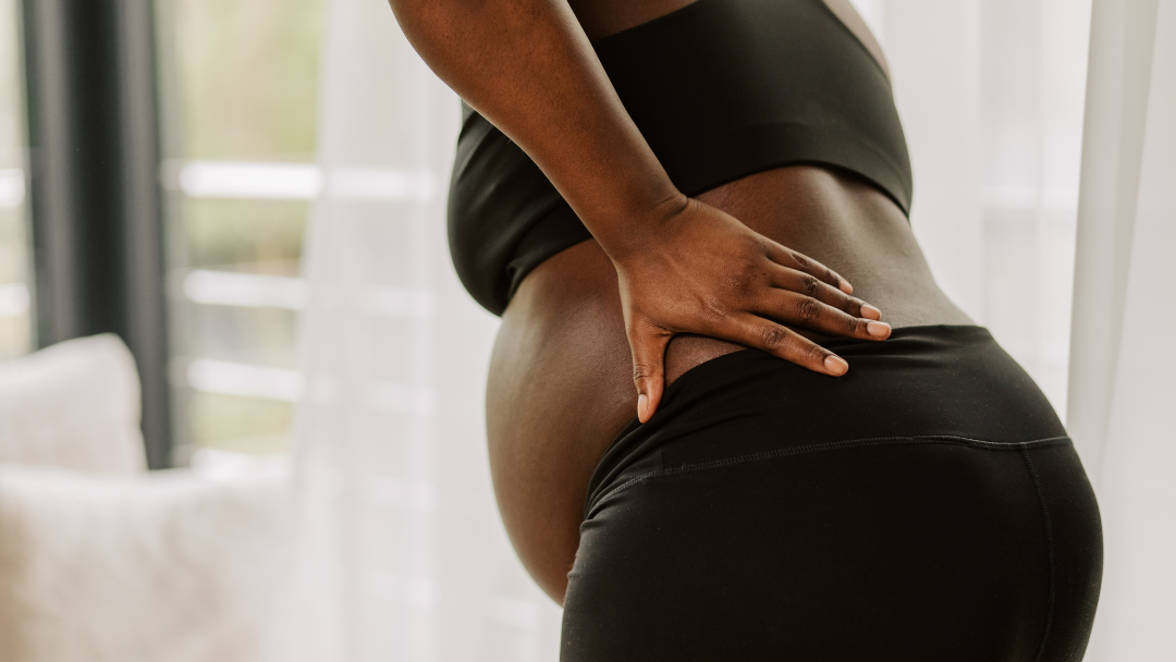 Black pregnant woman wearing black sports bra and black leggings hunched forward with hand on back to signify pregnancy pain.